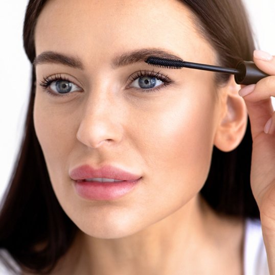 Portret kobiety w delikatnym makeupie malującej przed lustrem rzęsy tuszem