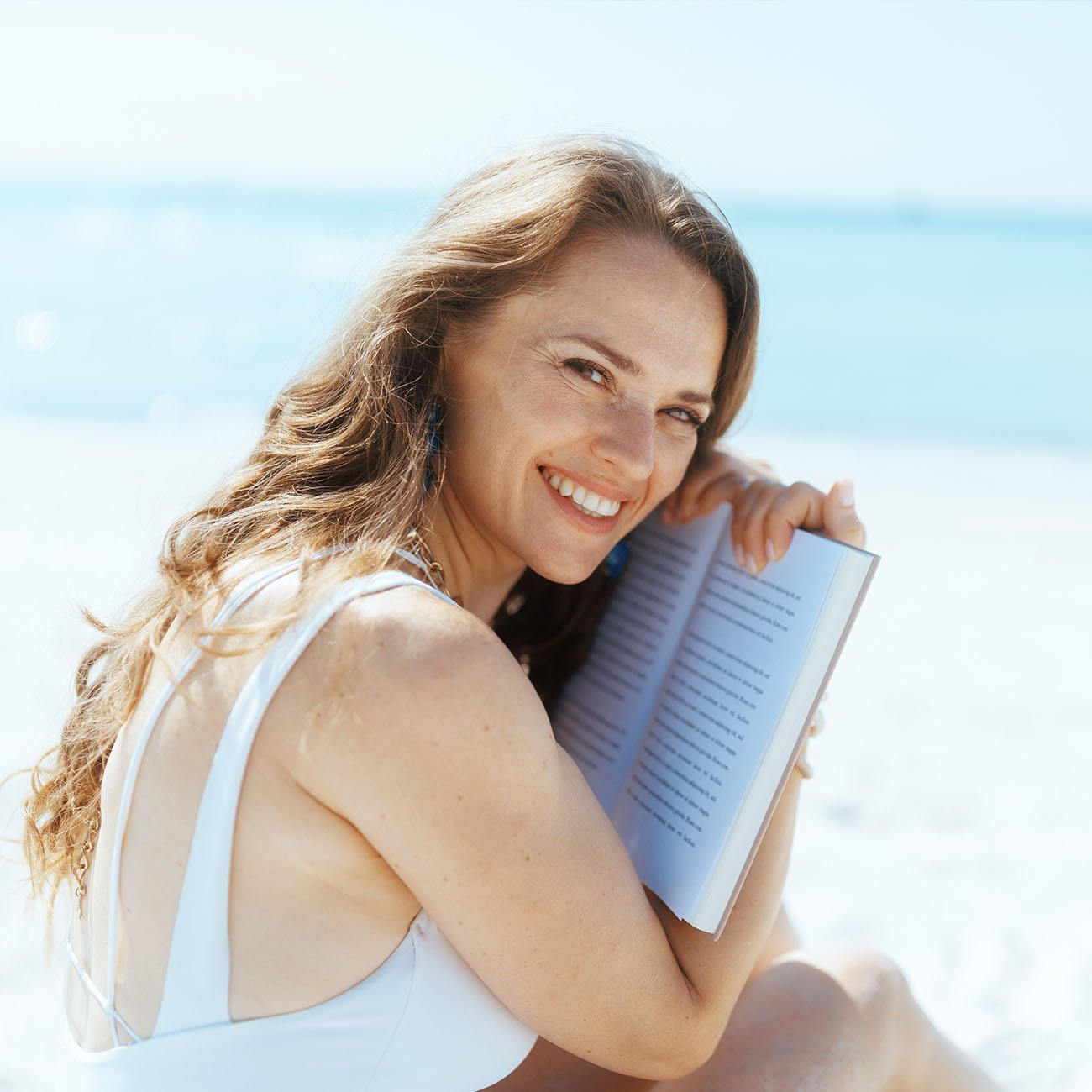 Uśmiechnięta kobieta w lekkim makijażu przytula otwartą książkę na plaży