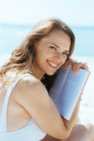 Uśmiechnięta kobieta w lekkim makijażu przytula otwartą książkę na plaży