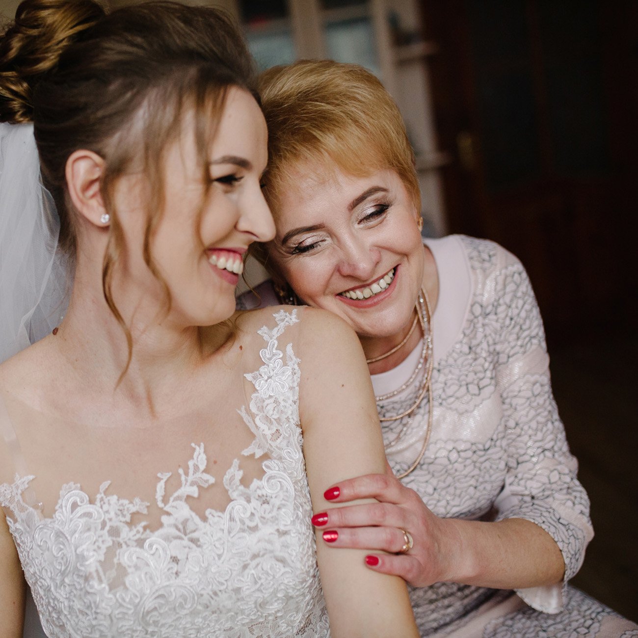 Mama siedzi na kanapie obok swojej córki w sukni ślubnej, przytula się do niej. Obie się uśmiechają, tworząc radosny portret rodziny.