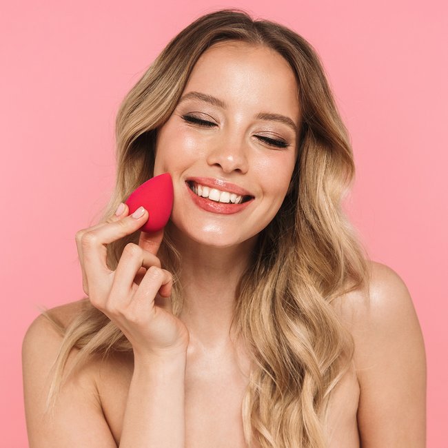 Portret młodej kobiety na różowym tle, nakładającej bazę pod makijaż przy pomocy beauty blendera