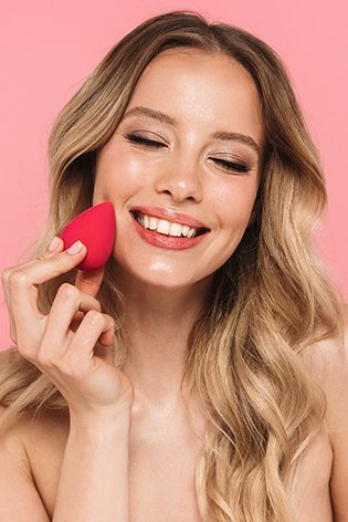 Portret młodej kobiety na różowym tle, nakładającej bazę pod makijaż przy pomocy beauty blendera