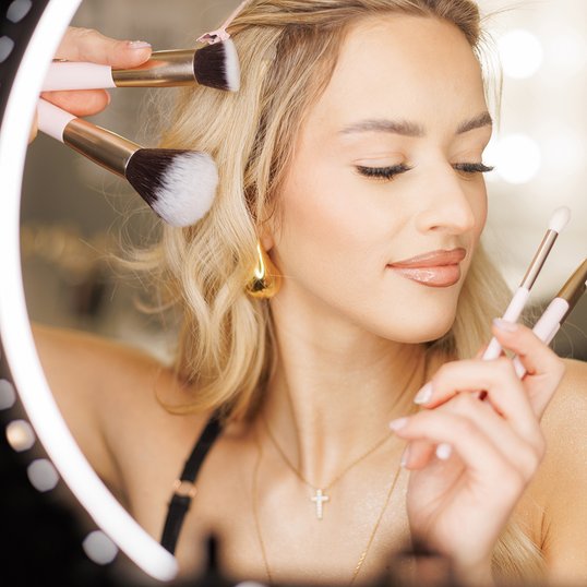 Blondwłosa kobieta przed lustrem trzyma w ręku pędzle do makijażu