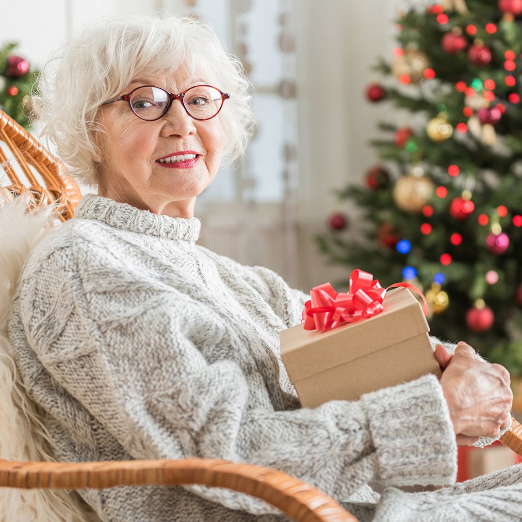 Uśmiechnięta seniorka na fotelu trzymająca świąteczny prezent, w tle choinka