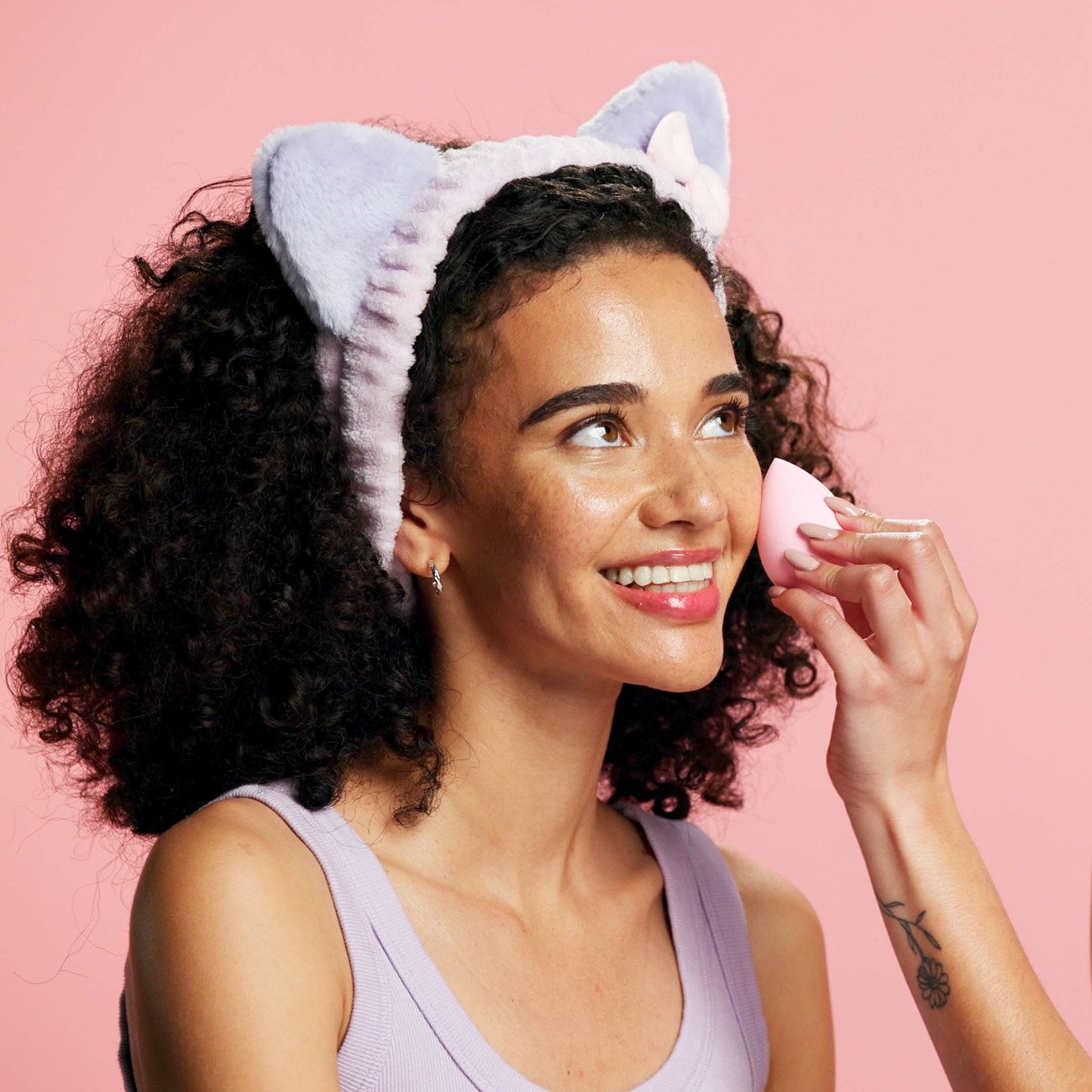 Dziewczyna na różowym tle, w opasce ochraniających włosy, druga dziewczyna nakłada jej na twarz podkład gąbeczką