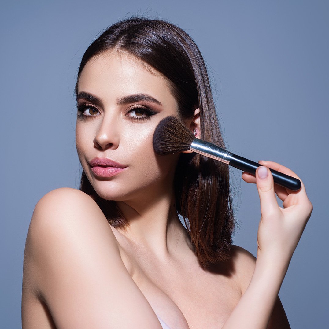 Młoda brunetka w mocnym makeupie na niebieskim tle, nakładająca kosmetyk pędzlem na twarz