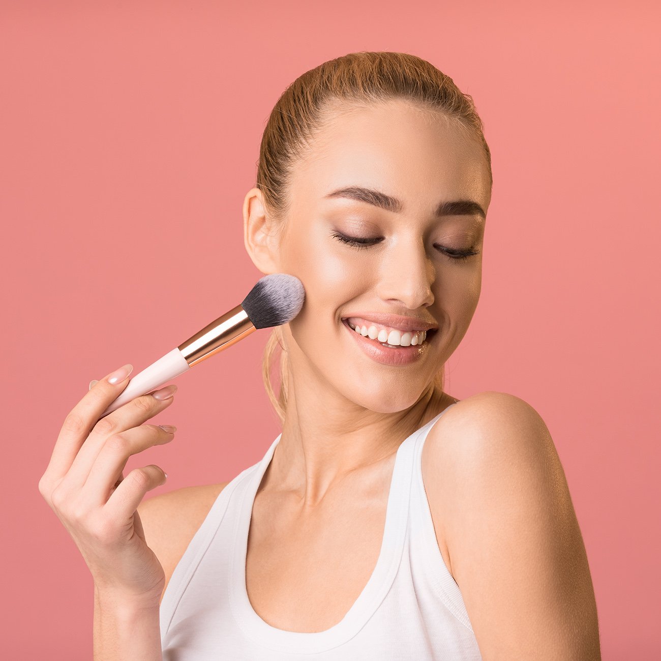 Młoda uśmiechnięta kobieta pozująca z pędzlem do makijażu na różowym tle